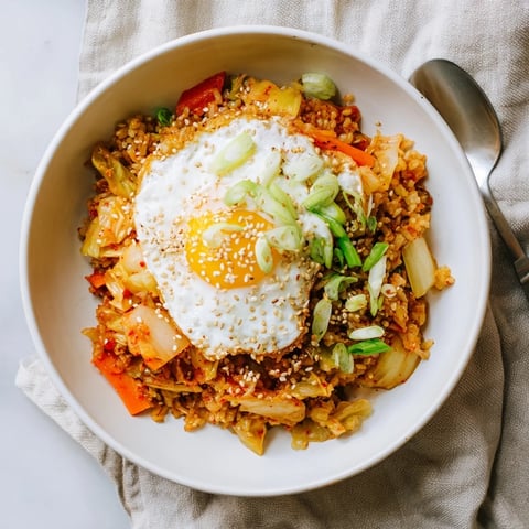 A steaming bowl of homemade Kimchi Fried Rice topped with a sunny-side-up fried egg and sesame seeds, served alongside crisp cucumber slices.  